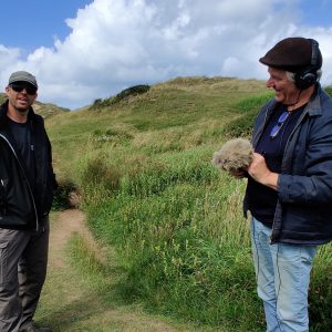 Two smiling men stand in the sand dunes, one holding a microphone and recording device