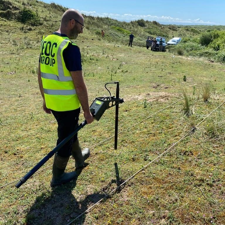 A man in a high vis jacket walks over a grassy dune with earth monitoring equipment